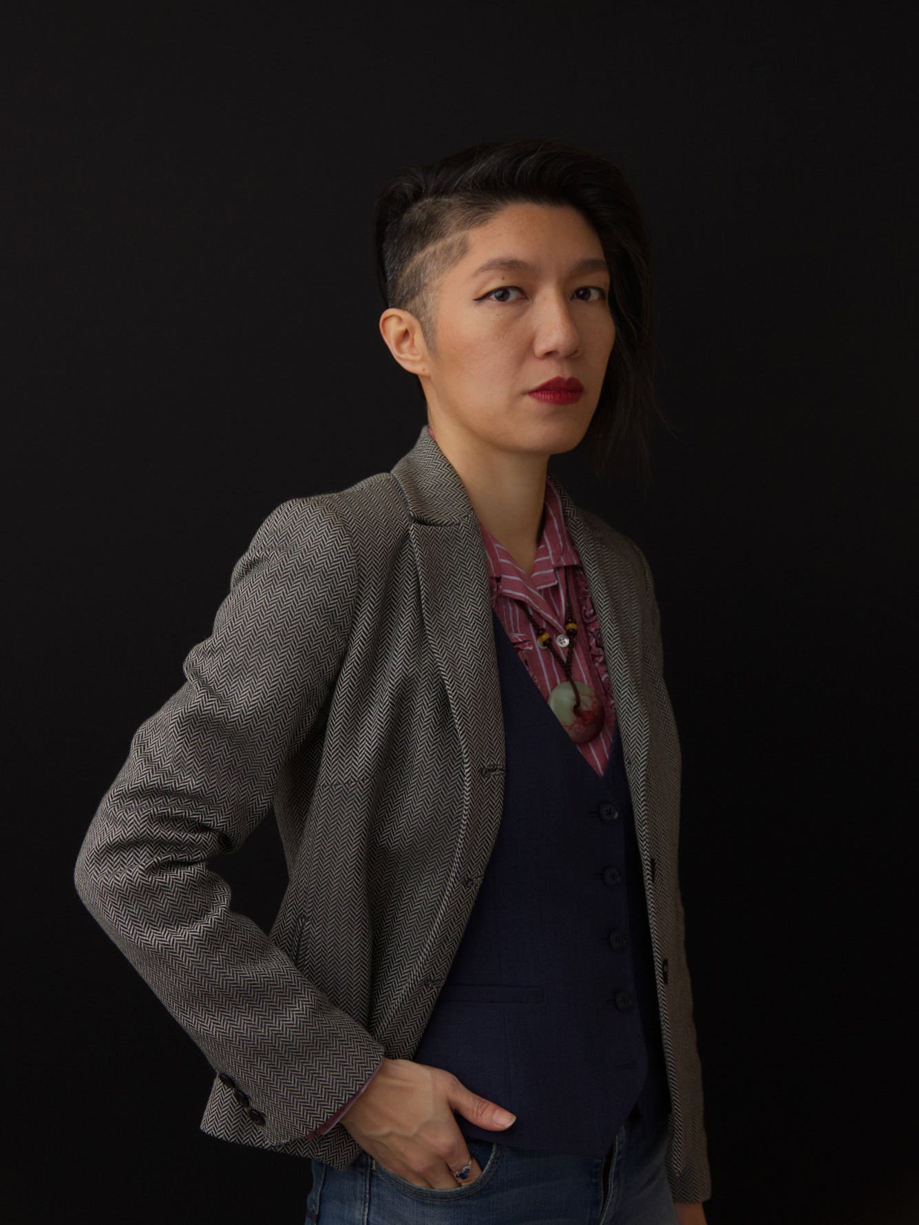 Jenie Gao portrait. Jenie is a young Asian femme and has short black hair with an undercut. They are wearing a grey herringbone blazer, a deep blue vest, a pink pinstriped buttondown shirt, and a circular red-green jade pendant. They are standing with their right hand tucked in their pocket.